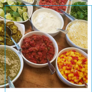 A variety of small dishes filled with taco toppings including: corn salsa, sour cream, shredded cheese and limes